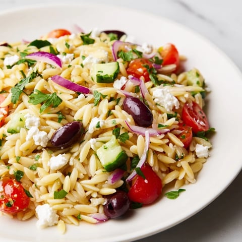 A close-up of Greek Orzo Salad with cherry tomatoes, cucumber, and crumbled feta on a rustic table.  