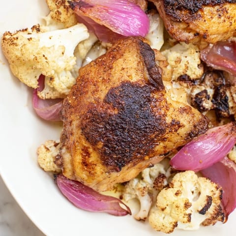 A close-up of seasoned chicken thighs and tender cauliflower florets roasted on a sheet pan, ready to serve.  